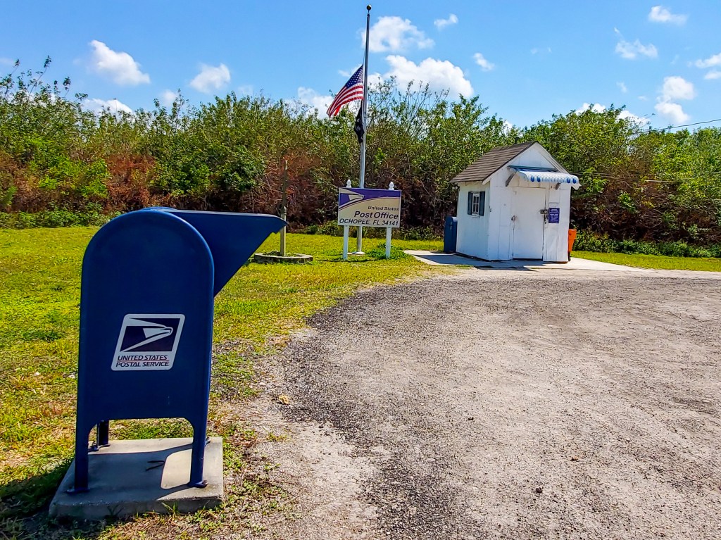 Post office box sits in from of very small building with sign with words Post Office Ochopee, FL